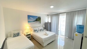 A hotel room with two beds, a desk, a lamp, a water dispenser, and a painting of a beach scene. - CARIBBEAN STUDIOS POOL BEACH & SPA  (Punta Cana)