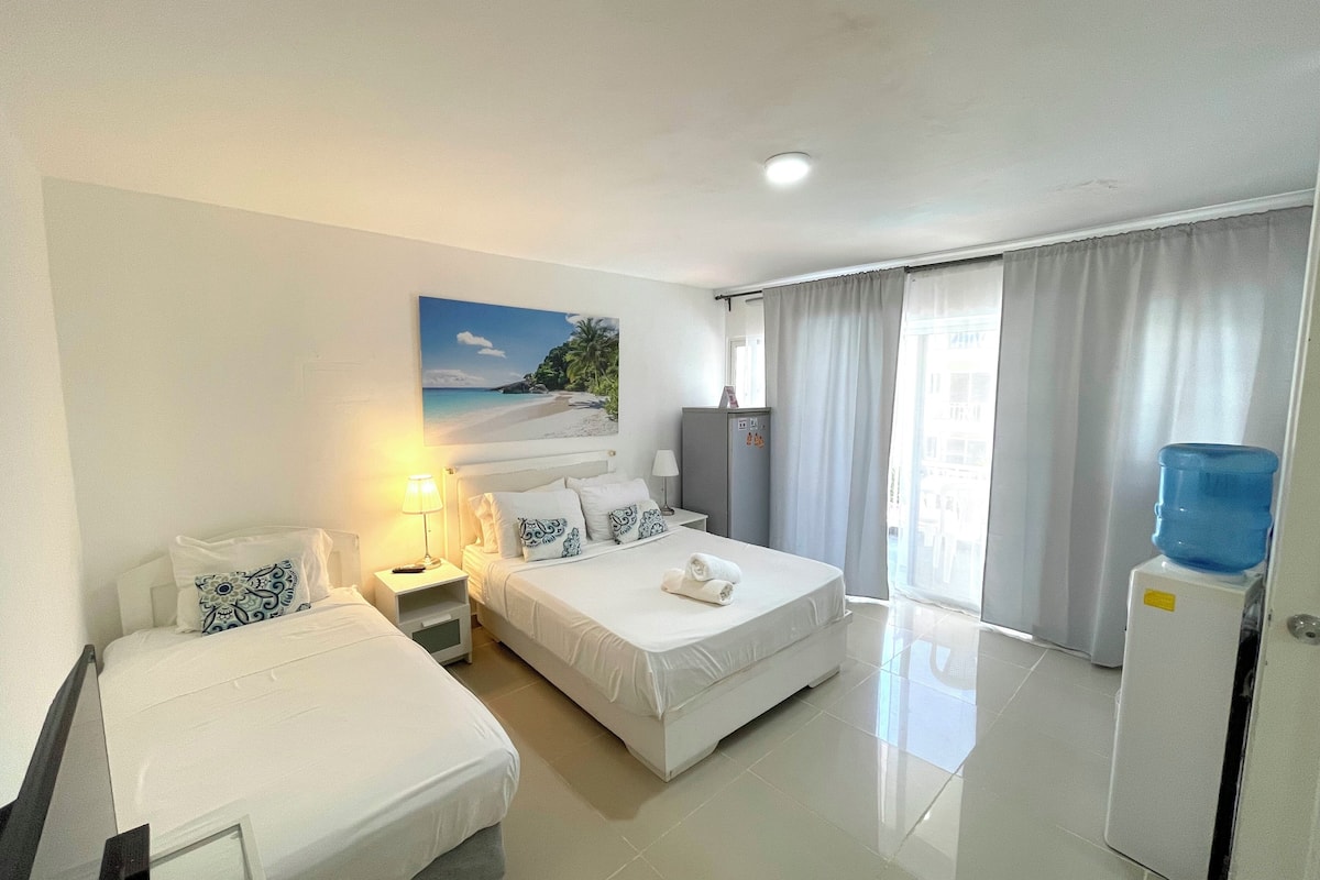 Habitación de hotel con dos camas, un escritorio, una lámpara, un dispensador de agua y un cuadro de una playa.