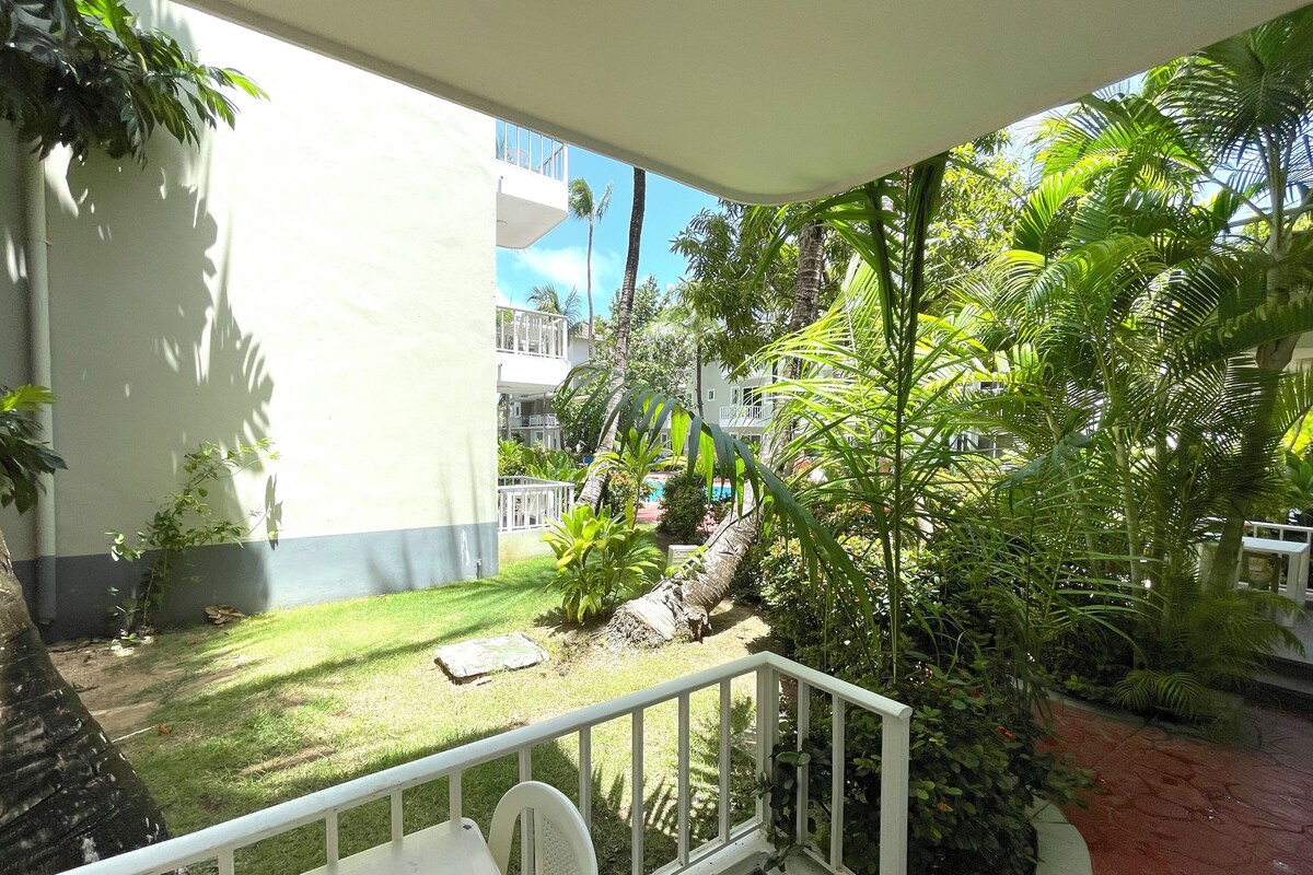 A balcony with a white railing, a small table, and a chair. There is a grassy area with plants and a palm tree.