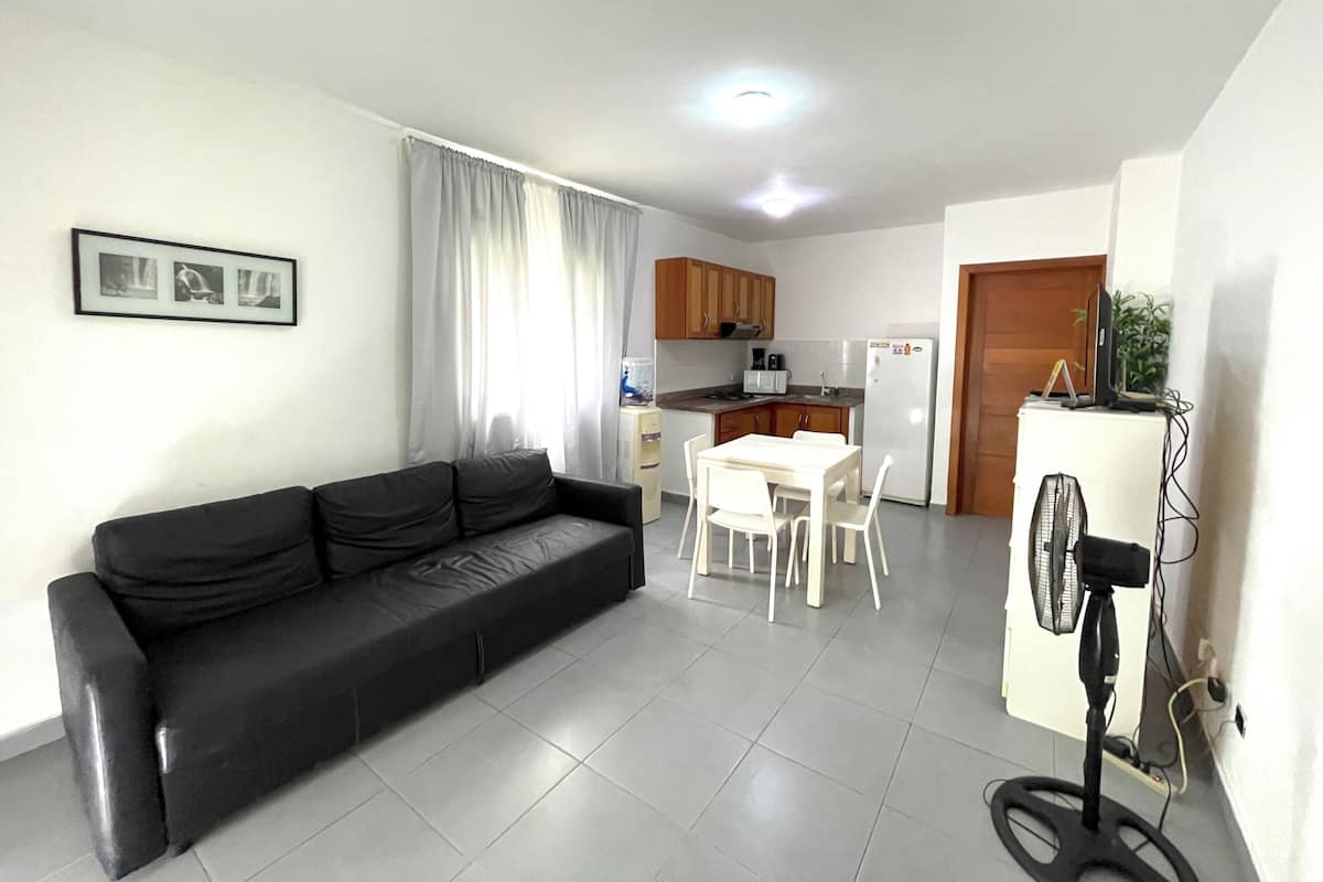 Un salón con un sofá negro, una mesa de comedor con sillas blancas, una zona de cocina con armarios de madera, un refrigerador blanco y un ventilador de pie.