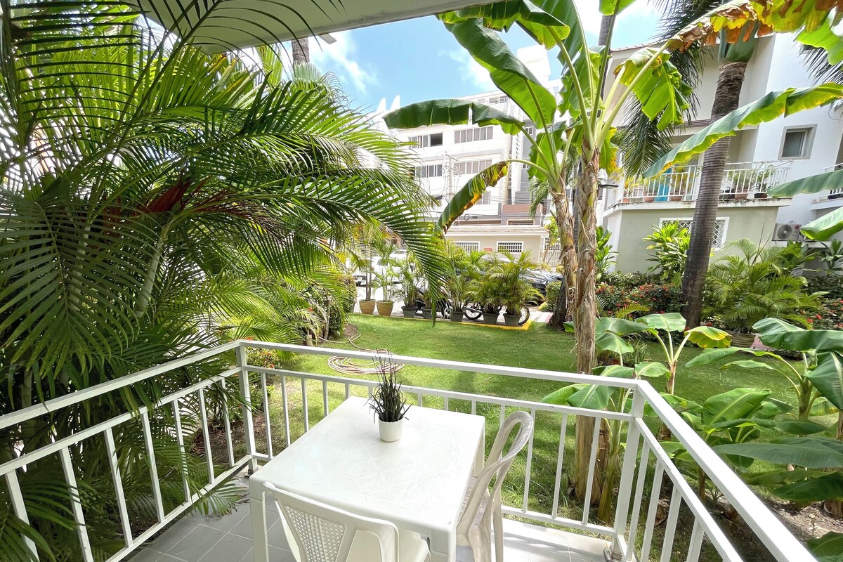 A balcony with a white table and chairs, surrounded by lush greenery and a view of a well-maintained garden and buildings in the background.