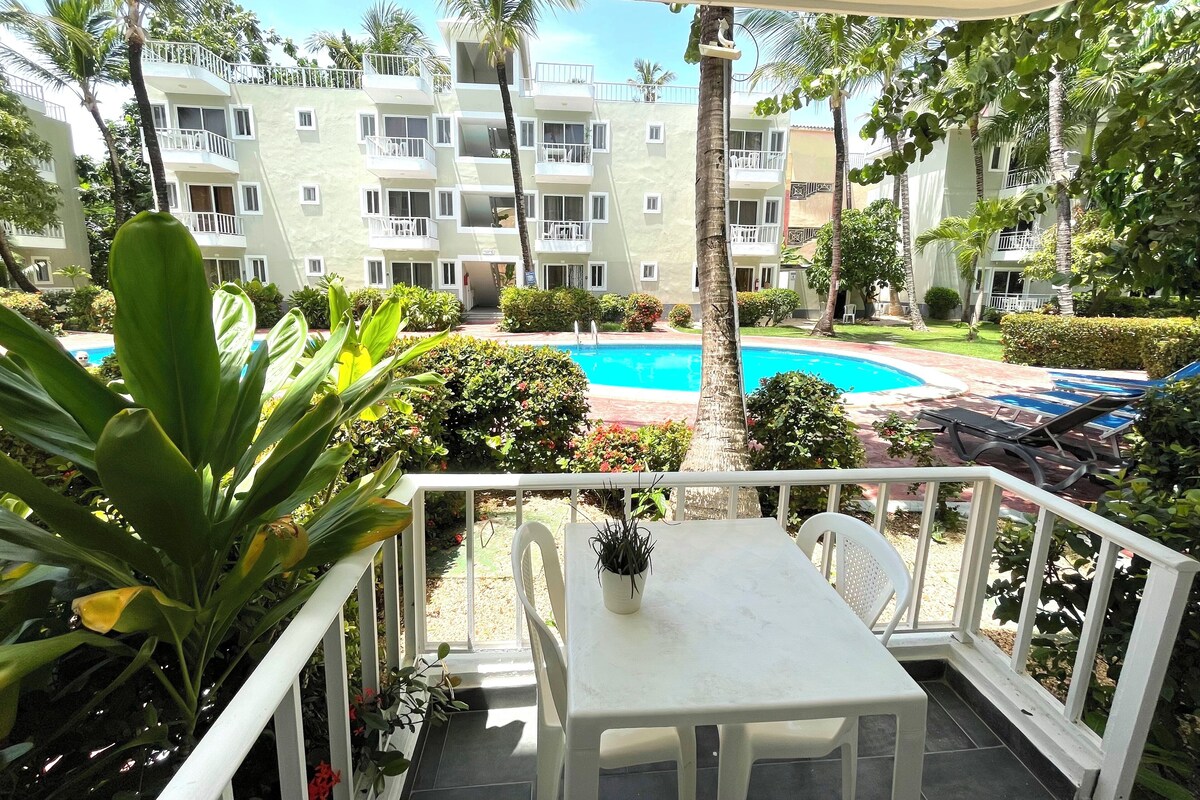A balcony with a view of a swimming pool, surrounded by lush greenery and a multi-story building with balconies.