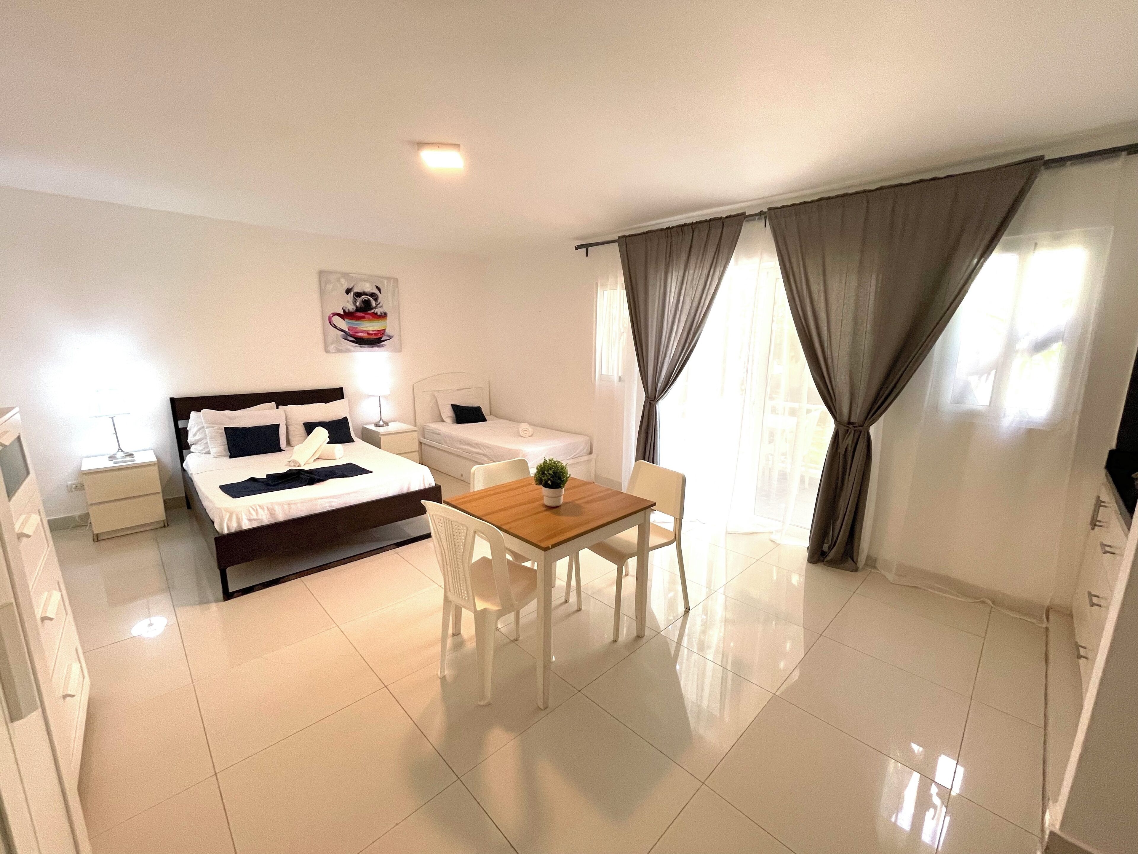 A hotel room with a bed, a desk, a chair, a small table, a nightstand, and a window with curtains.