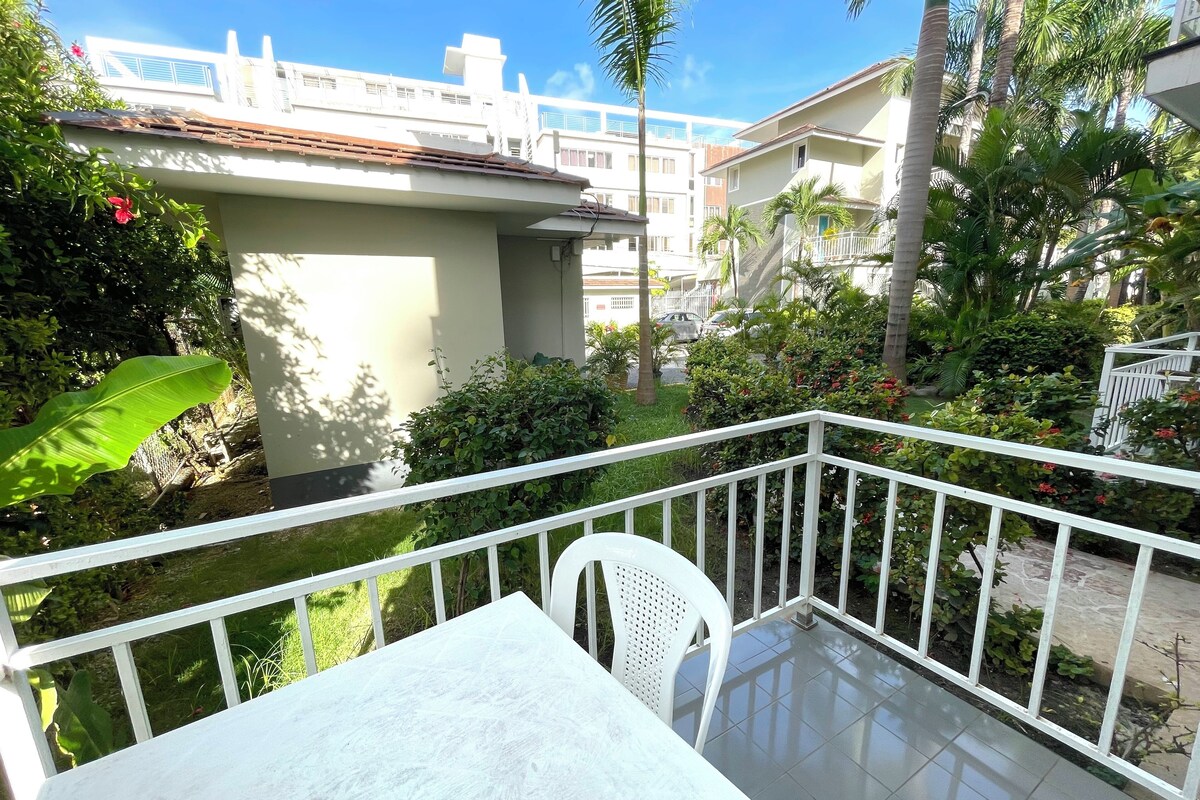 A balcony with a white table and chair, surrounded by lush greenery and a white railing.