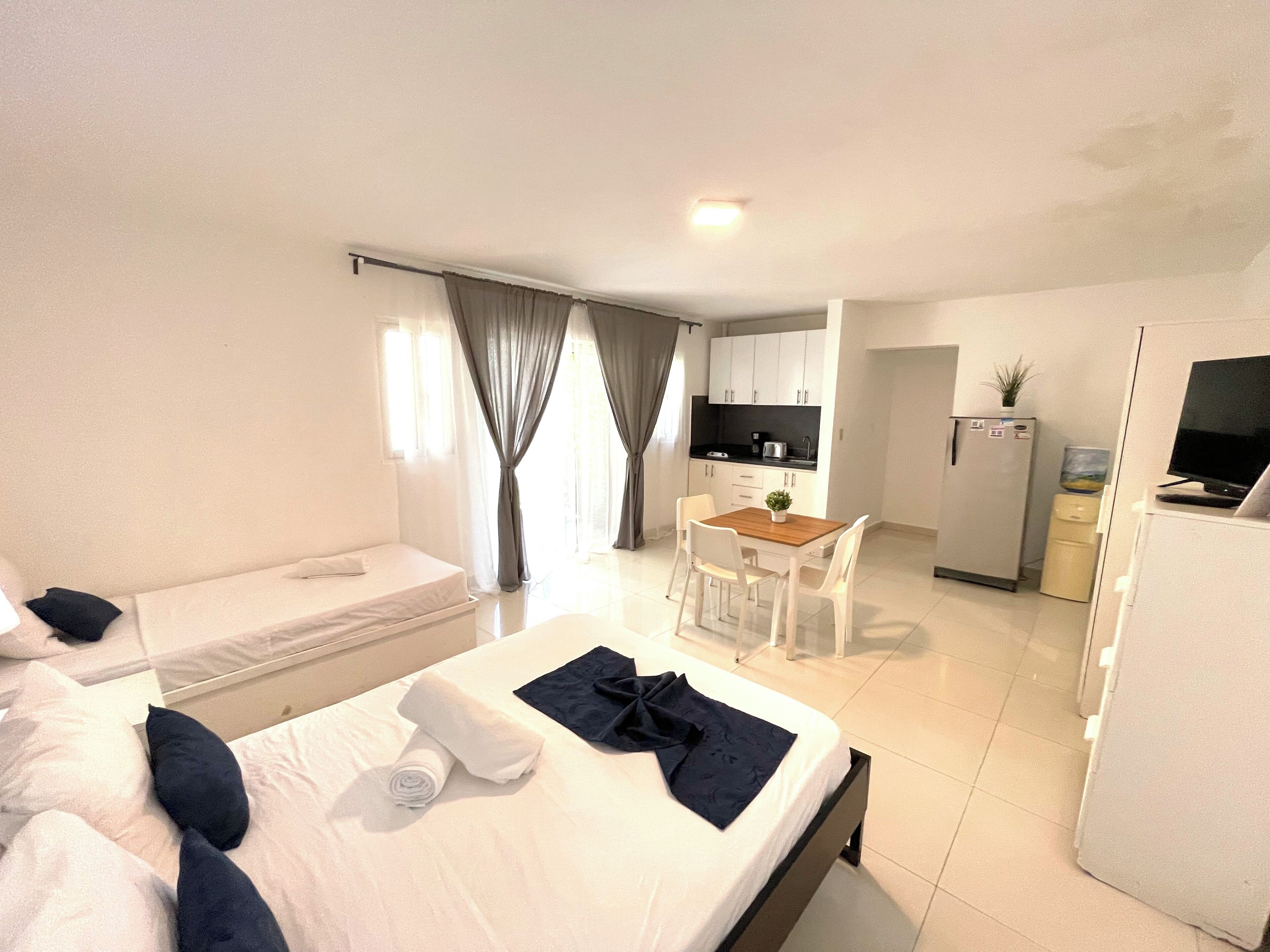A hotel room with a bed, a desk, a chair, a small kitchen area, and a TV.