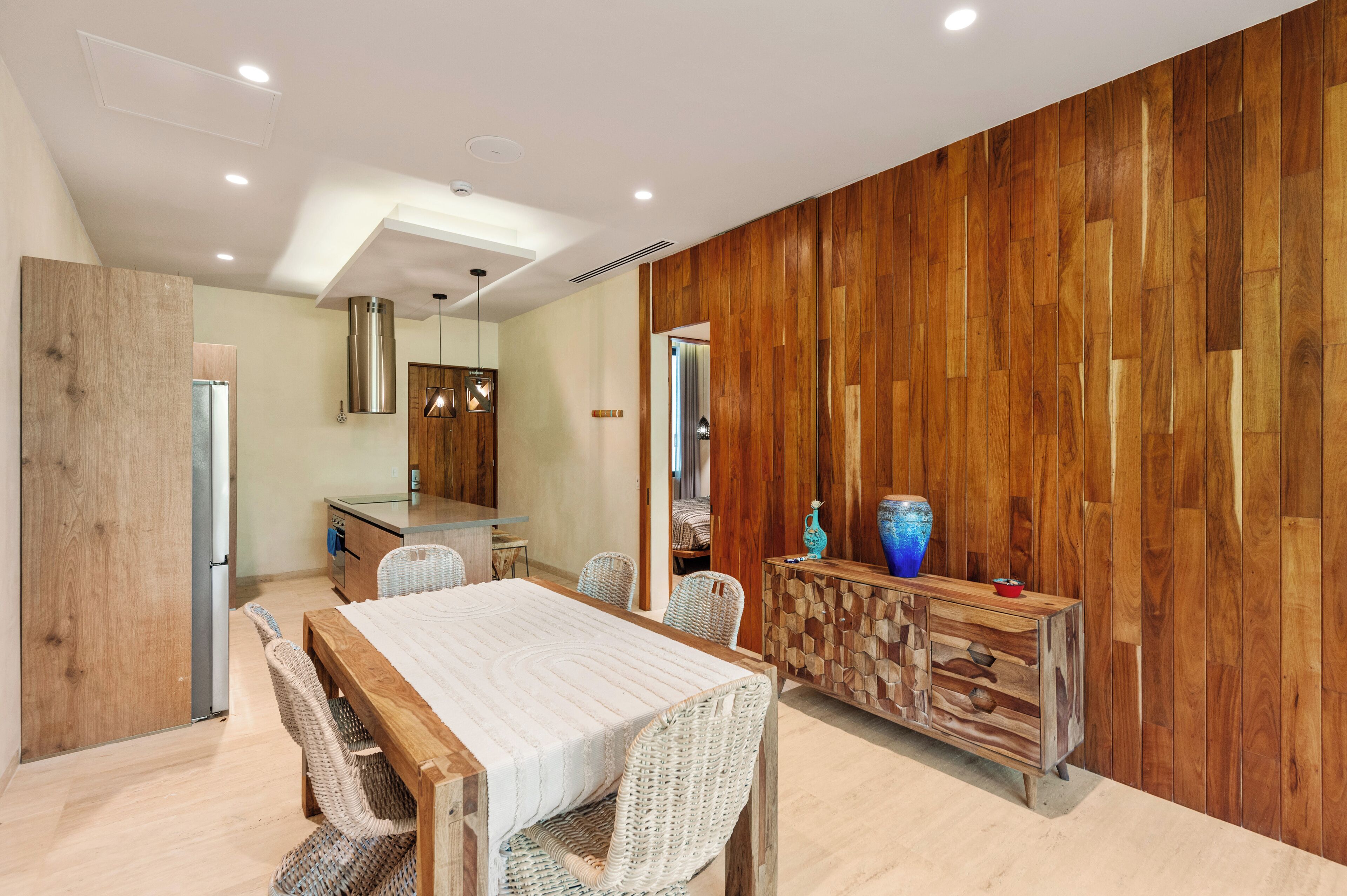 A modern dining area with a wooden table and chairs, a kitchen in the background, and a wooden panel wall.