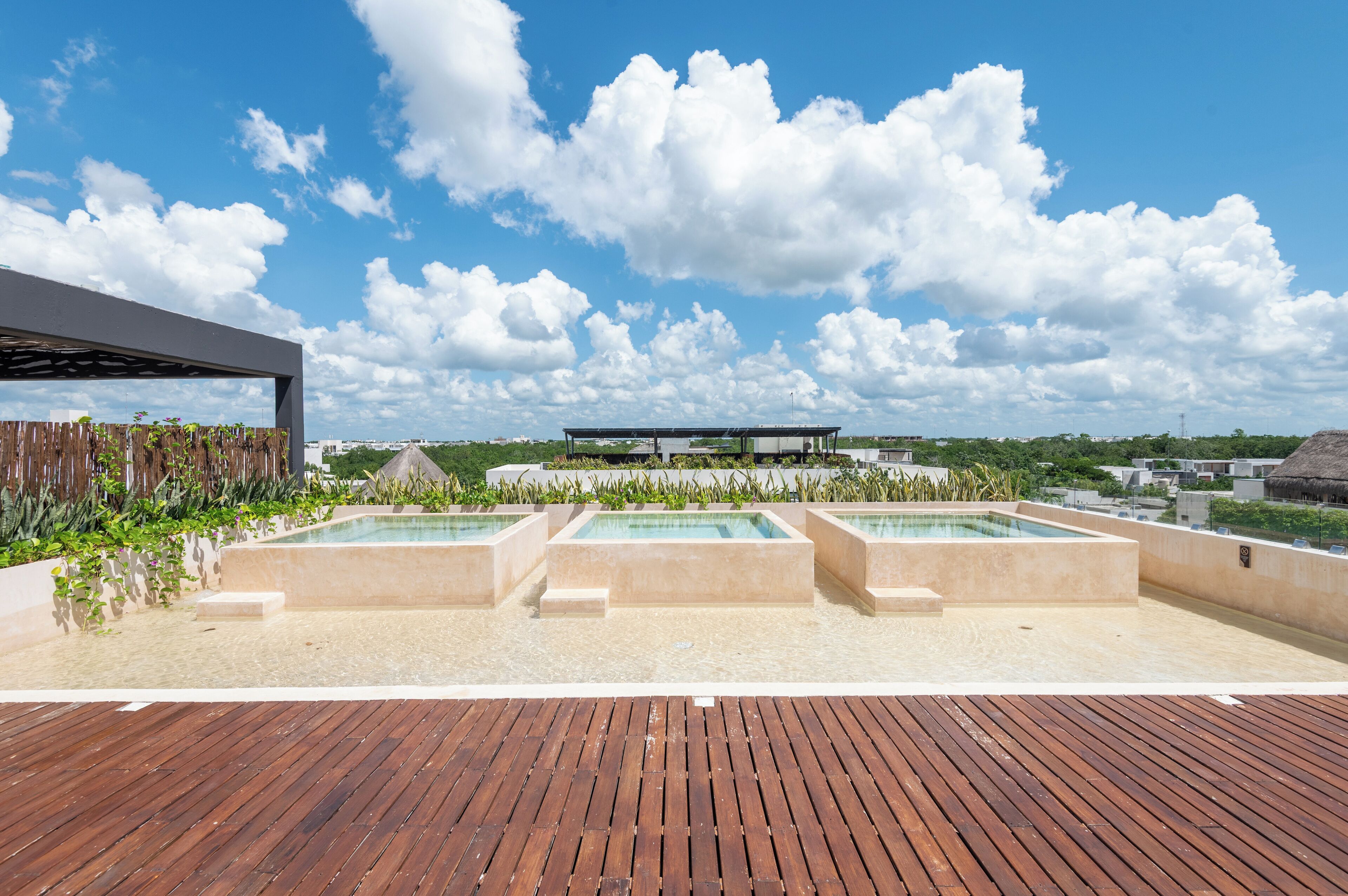 A rooftop pool area with three rectangular pools, a wooden deck, and a modern building structure in the background.
