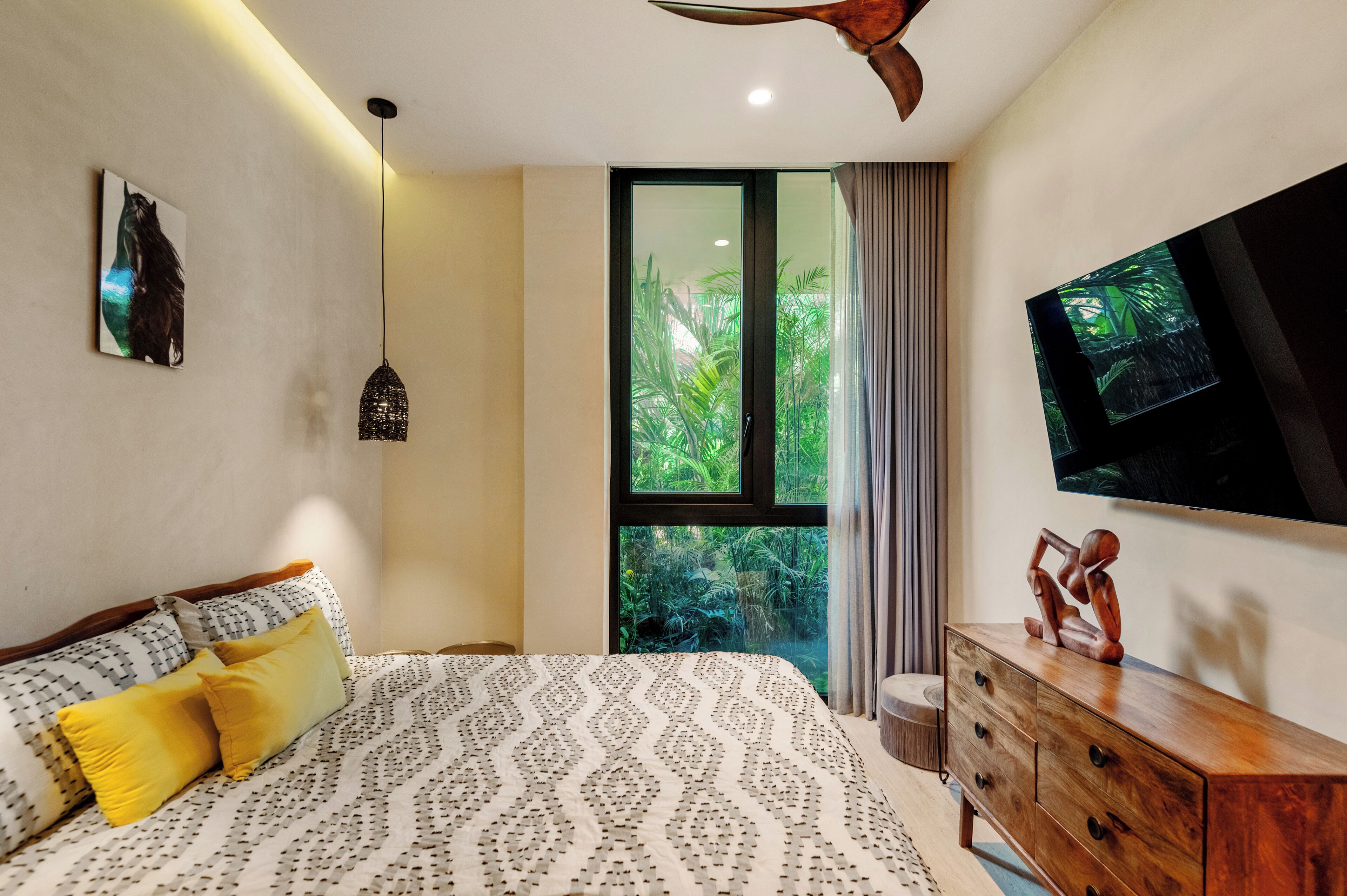 A bedroom with a bed, a TV, a wooden dresser, and a window with a view of greenery.