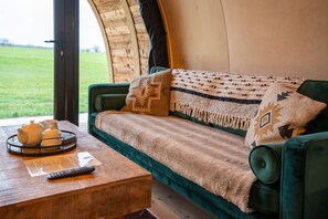 Ein gemütliches Zimmer mit einem grünen Sofa, einem Holztisch mit einer Teewanne und einer Fernbedienung, und Blick auf ein grünes Feld durch ein großes Fenster.