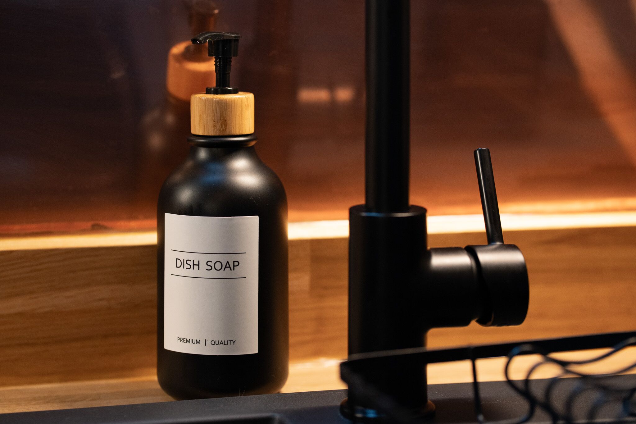 A black bottle labeled "Dish Soap" with a wooden cap and a pump dispenser, placed on a wooden surface next to a black object, possibly a camera or microphone, in a well-lit indoor setting.