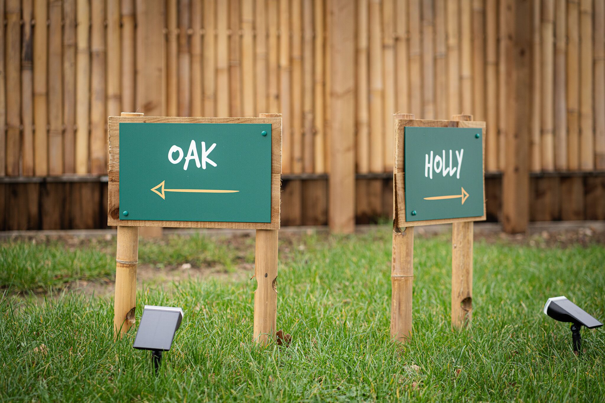 Two directional signs labeled "OAK" and "HOLLY" on a grassy area with a wooden fence in the background.