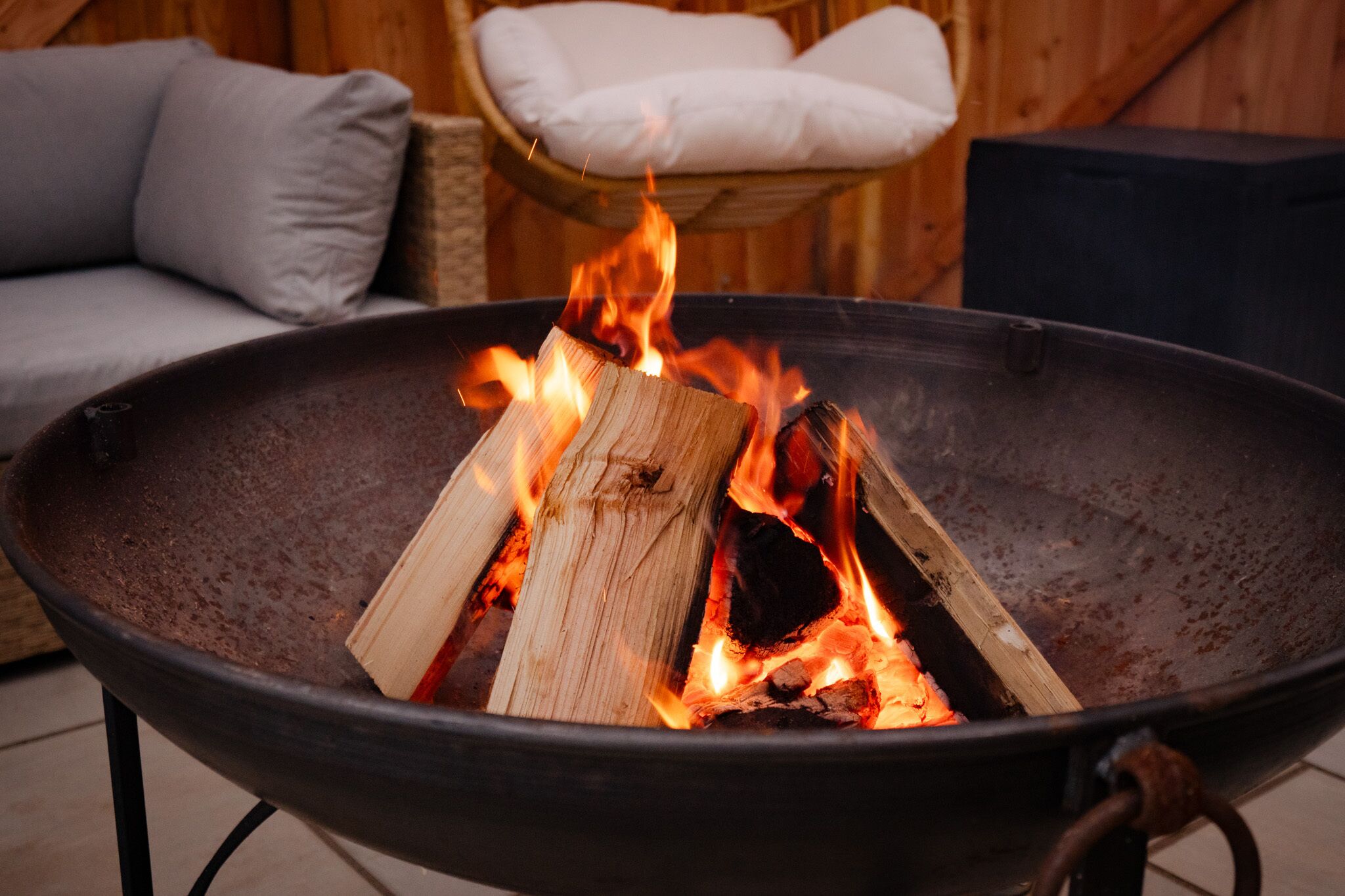 A fire pit with burning wood logs, a wicker chair with a white cushion, and a grey sofa in the background.