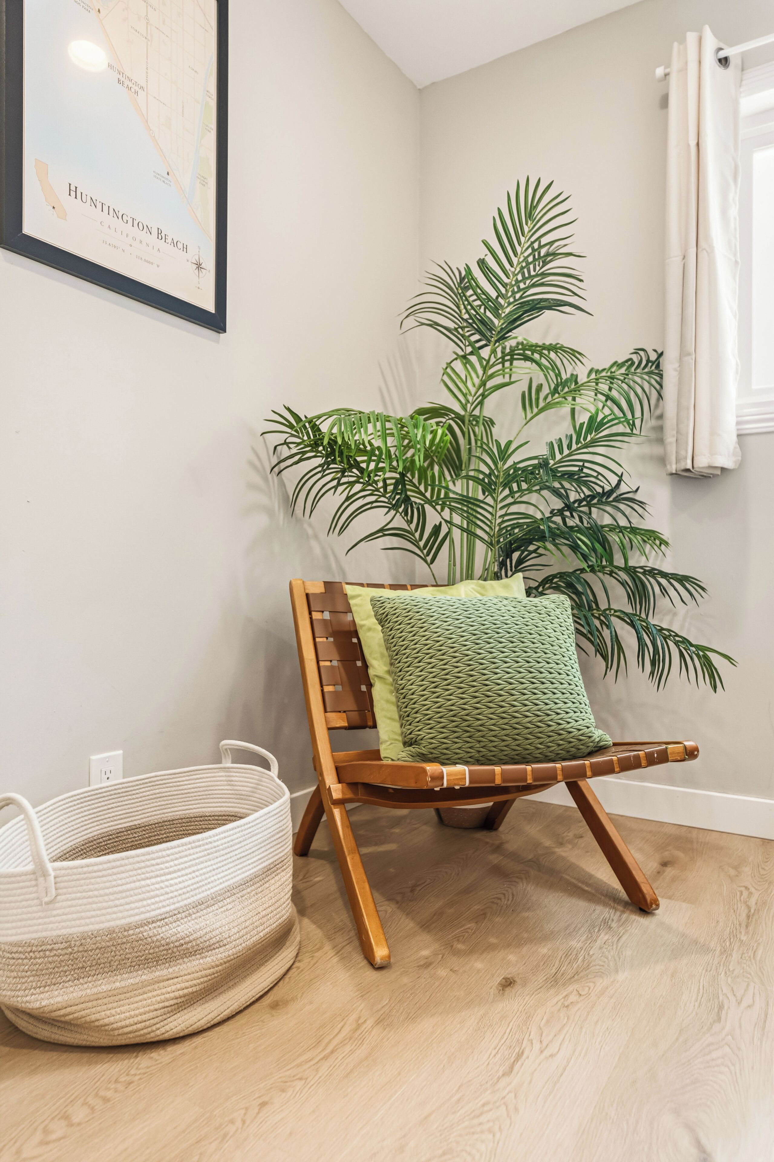 Une pièce d’angle avec une chaise en bois, un panier en osier, une grande plante en pot et une carte encadrée accrochée au mur.