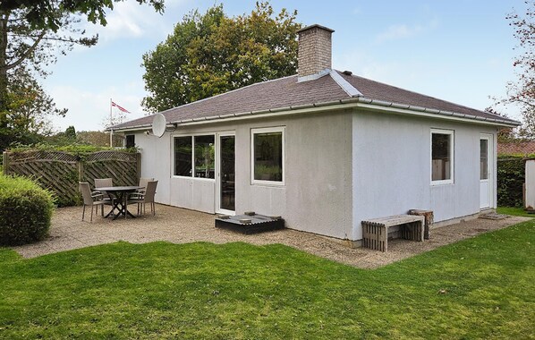 Une petite maison de plain-pied, avec un toit en tuiles, une cheminée et un patio comprenant une table et des chaises.