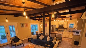 A spacious living area with a wooden ceiling, a dining table, sofas, and a kitchen in the background.