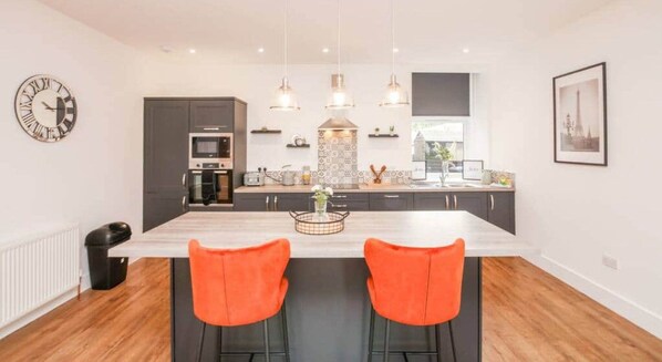 Una cocina moderna con una isla central, luces colgantes y un comedor con dos sillas naranjas.