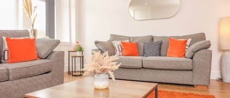 A modern living room with two grey sofas, orange cushions, a wooden coffee table, and a large mirror on the wall.