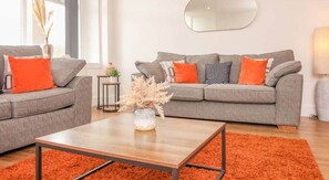 Un salon moderne avec deux canapés gris, des coussins orange, une table basse en bois et un grand miroir au mur.