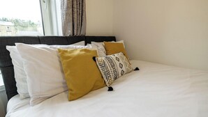 A neatly made bed with white and mustard pillows, a dark headboard, and a window with curtains. - Riverside - sleeps 6 guests  in 2 bedrooms (Hawick)