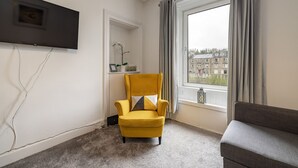 A room with a yellow armchair, a grey sofa, a flat-screen TV mounted on the wall, a window with curtains, and a view of buildings outside. - Riverside - sleeps 6 guests  in 2 bedrooms (Hawick)