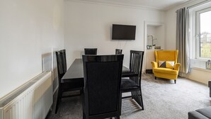 A modern room with a dining table and chairs, a flat-screen TV mounted on the wall, a yellow armchair, and a window with curtains. - Riverside - sleeps 6 guests  in 2 bedrooms (Hawick)