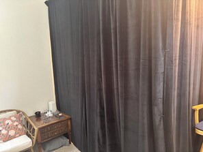 A room with a dark curtain, a wooden nightstand with a lamp, and a chair.