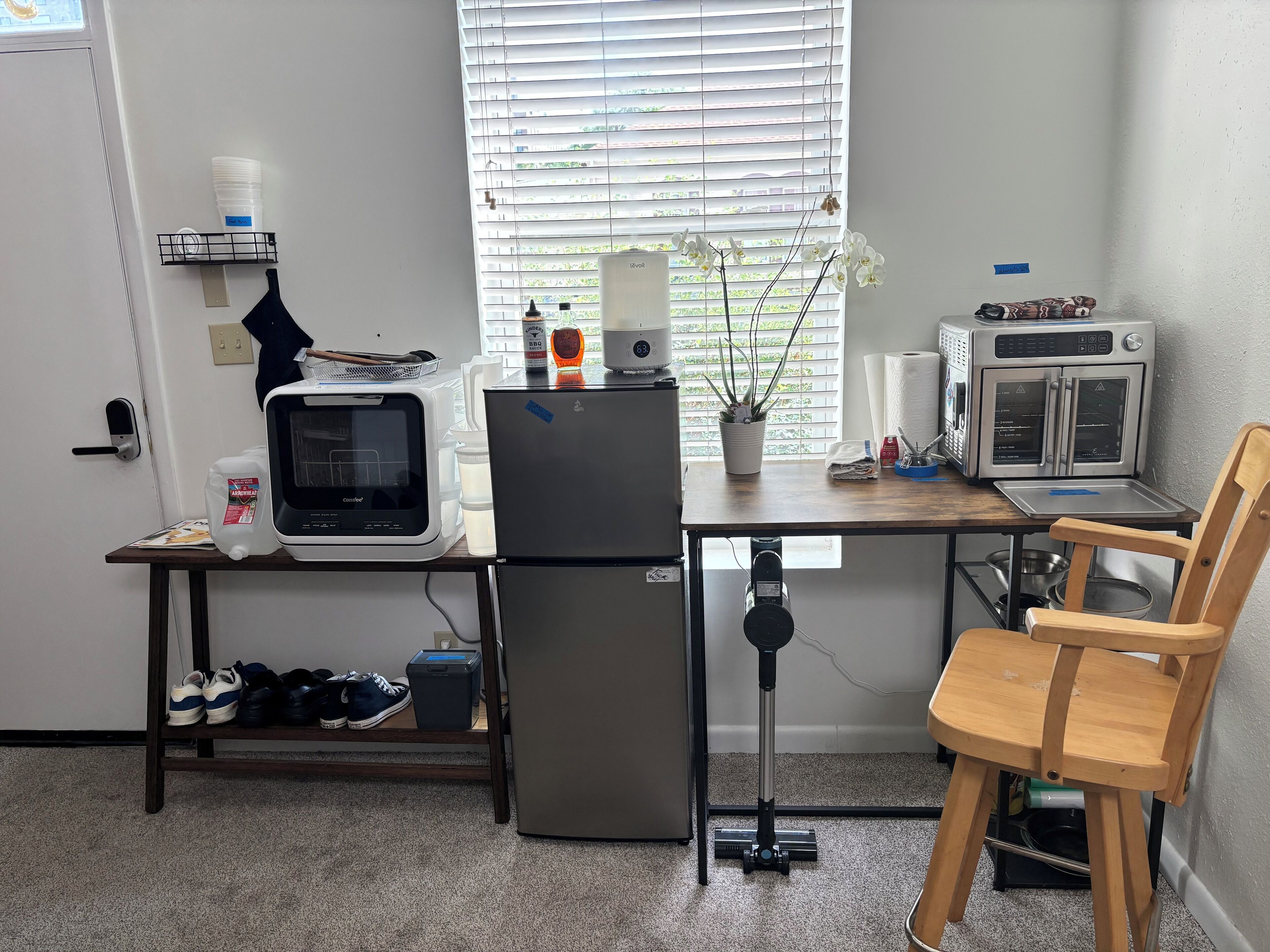 A compact kitchenette with a microwave, refrigerator, and oven. A small table with a chair is positioned near the window, which has blinds.