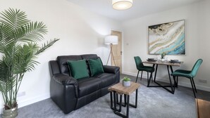A modern living room with a black leather sofa, green cushions, a small wooden coffee table, a dining table with chairs, a large abstract painting, and a tall floor lamp.