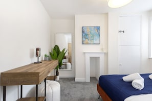 Un dormitorio con un escritorio de madera, una cama con colcha azul, una puerta blanca y un cuadro en la pared.
