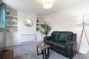 Ein modernes Wohnzimmer mit einem schwarzen Ledersofa, einem Holztisch und einer Stehlampe. An der Wand befindet sich ein Heizkörper und ein Blumentopf.