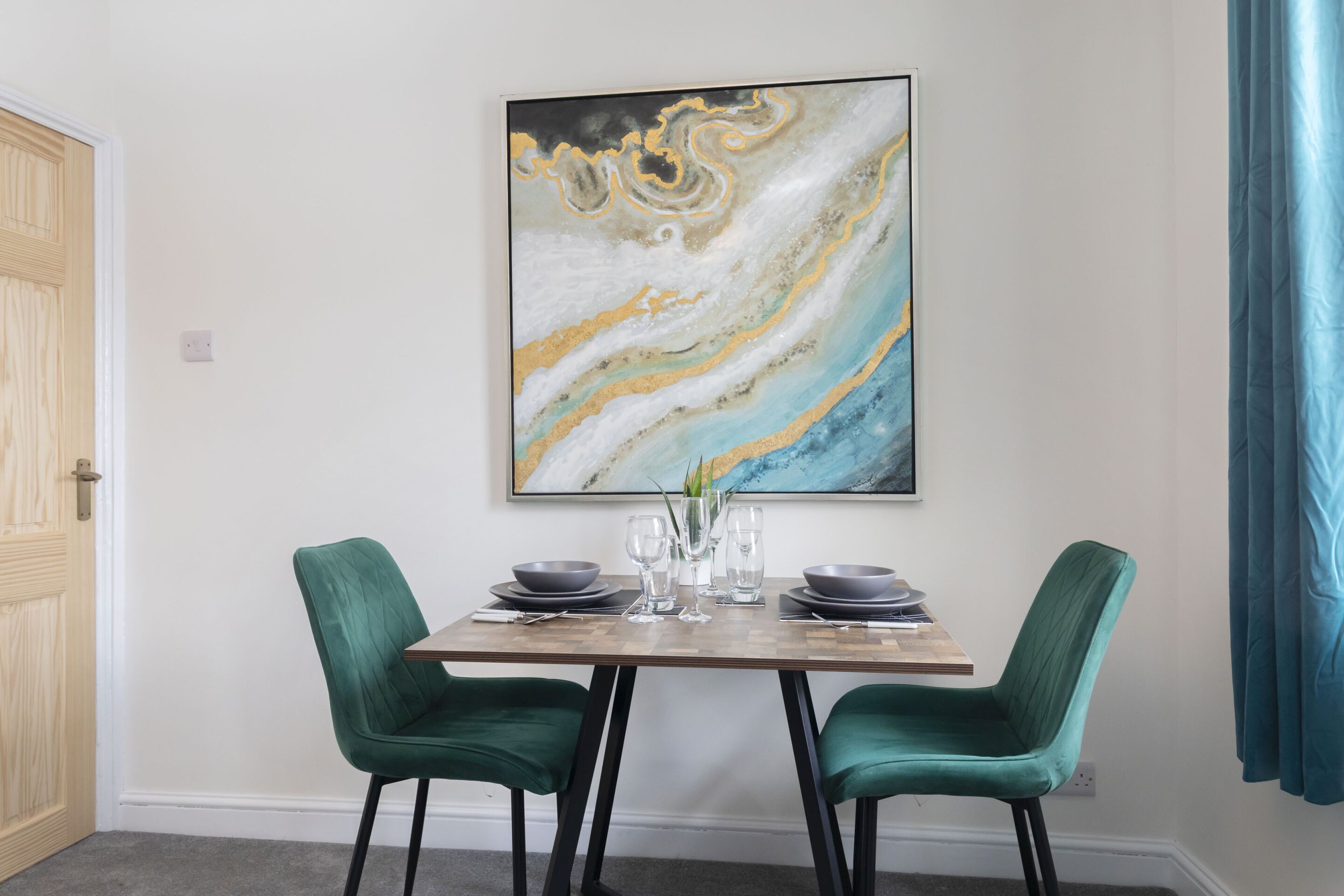 A dining area with a marble artwork, two green upholstered chairs, a wooden table, and a blue curtain.