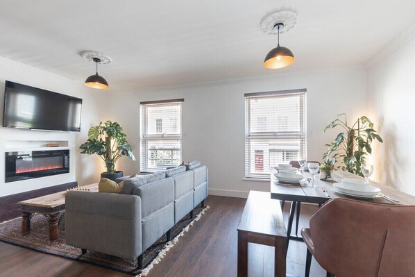 A modern living room with a grey sofa, a wooden coffee table, a fireplace, a TV mounted on the wall, and two windows with blinds.