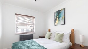 Quarto bem arrumado com cama, mesa de cabeceira, abajur, janela com persianas e um quadro na parede.