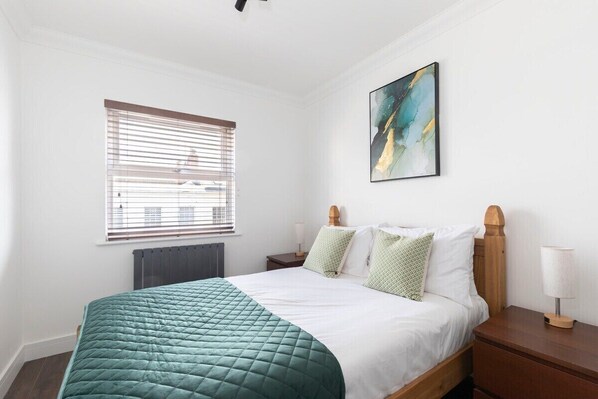 Un dormitorio ordenado con una cama, una mesita de noche, una lámpara, una ventana con persianas y un cuadro en la pared.