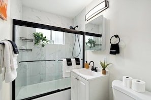 Une salle de bain moderne avec une douche à parois de verre, des carreaux de marbre blanc, un lavabo blanc avec un miroir au-dessus et des toilettes avec du papier toilette.