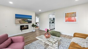Una sala de estar moderna con chimenea, un cuadro grande de playa y una cocina visible a través de una puerta abierta.