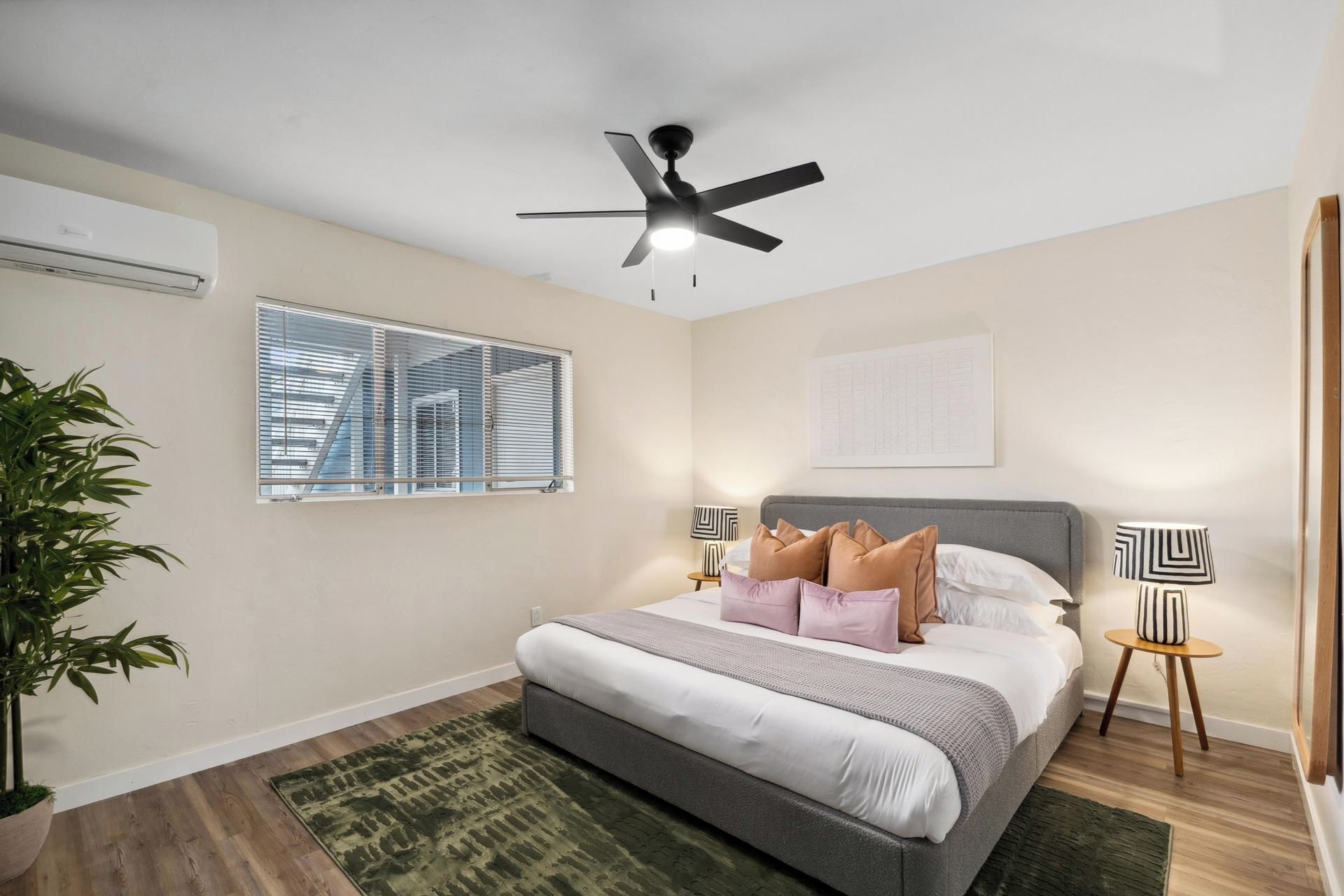 Une chambre à coucher comprenant un lit, un ventilateur de plafond, une fenêtre avec des stores, une table de chevet avec une lampe et une plante en pot.