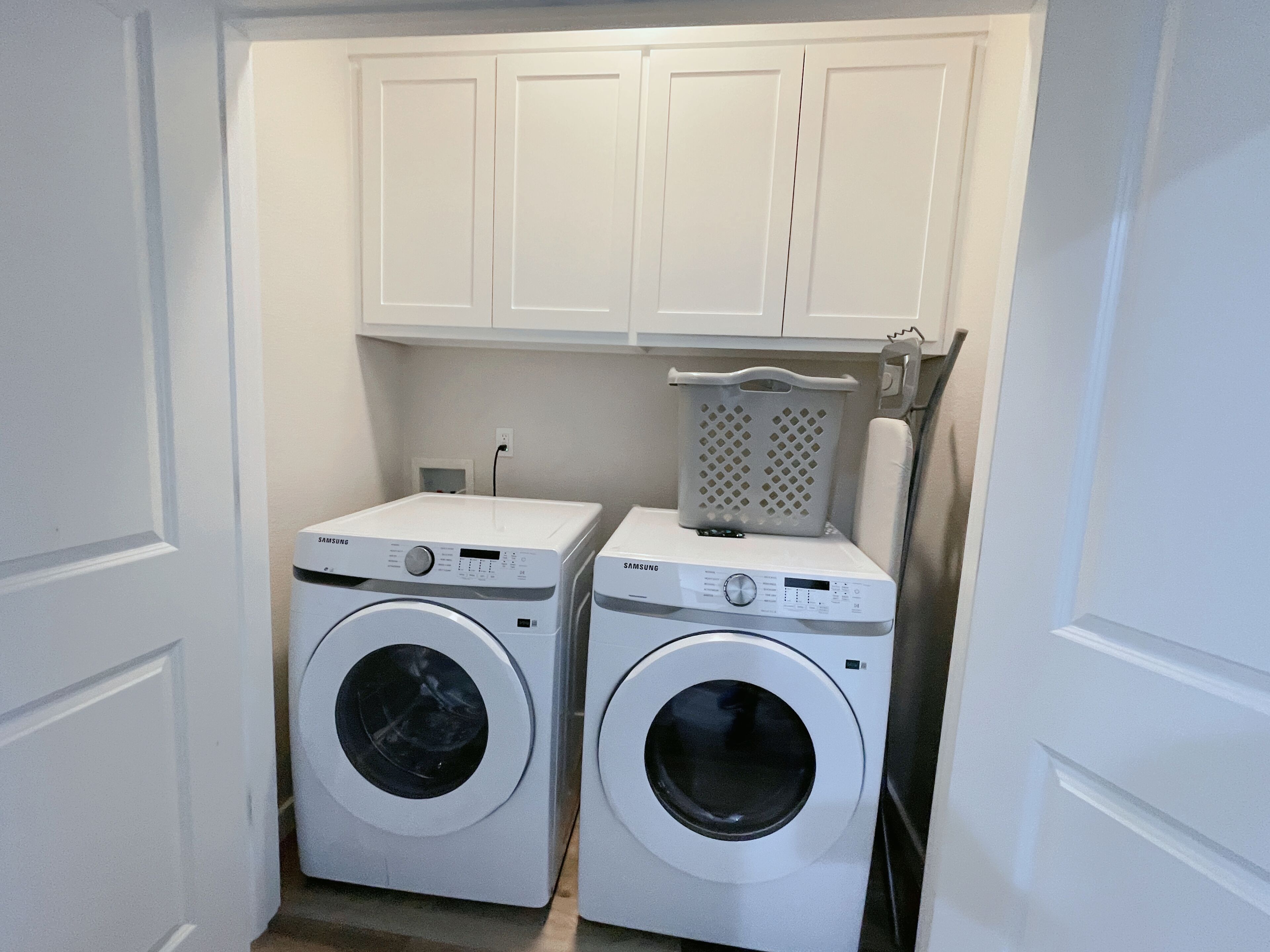 A laundry room with two white Samsung washing machines, a white laundry basket, and white cabinets above.