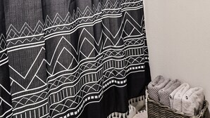 Un baño con cortina de ducha de estampado blanco y negro, una cesta de mimbre con toallas y una ventana con persianas.