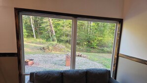 Un sofá gris orientado hacia una ventana grande con vistas a una zona boscosa y un sendero de grava.