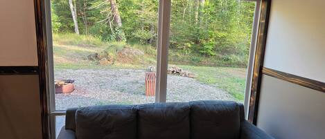A grey sofa facing a large window with a view of a forested area and a gravel path.