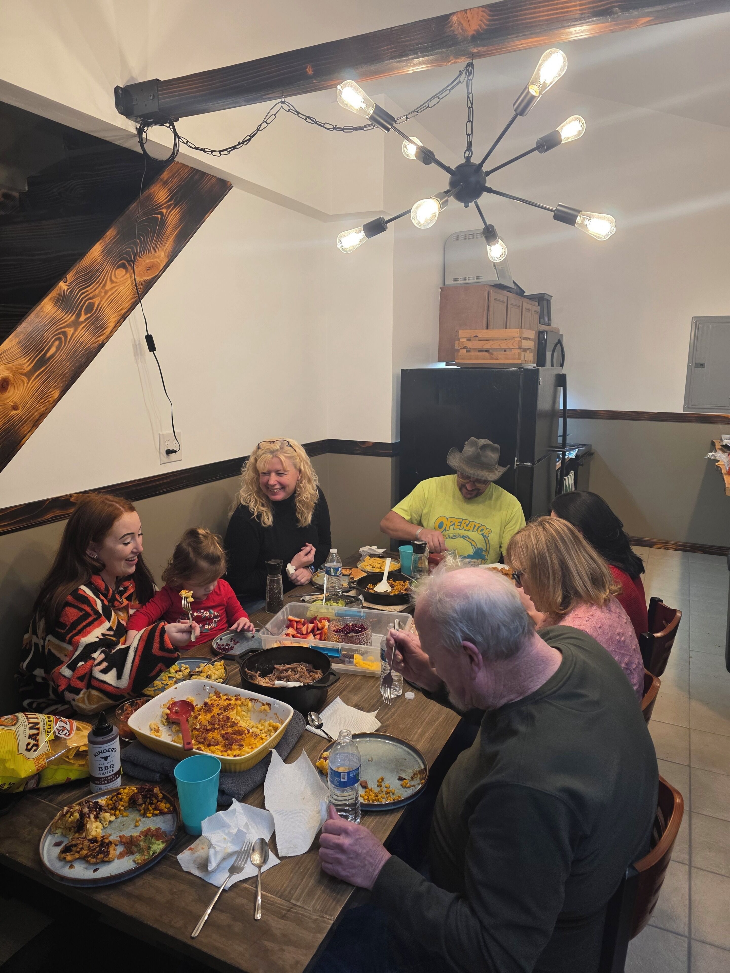 Un grupo de personas cenando en una mesa de madera con varios platos, un ventilador de techo y una cocina al fondo.