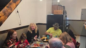 Un grupo de personas cenando en una mesa de madera con varios platos, un ventilador de techo y una cocina al fondo.