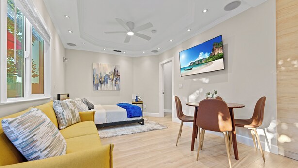 Een moderne woonkamer met een gele bank, een eettafel met stoelen, een plafondventilator en een televisie aan de muur.