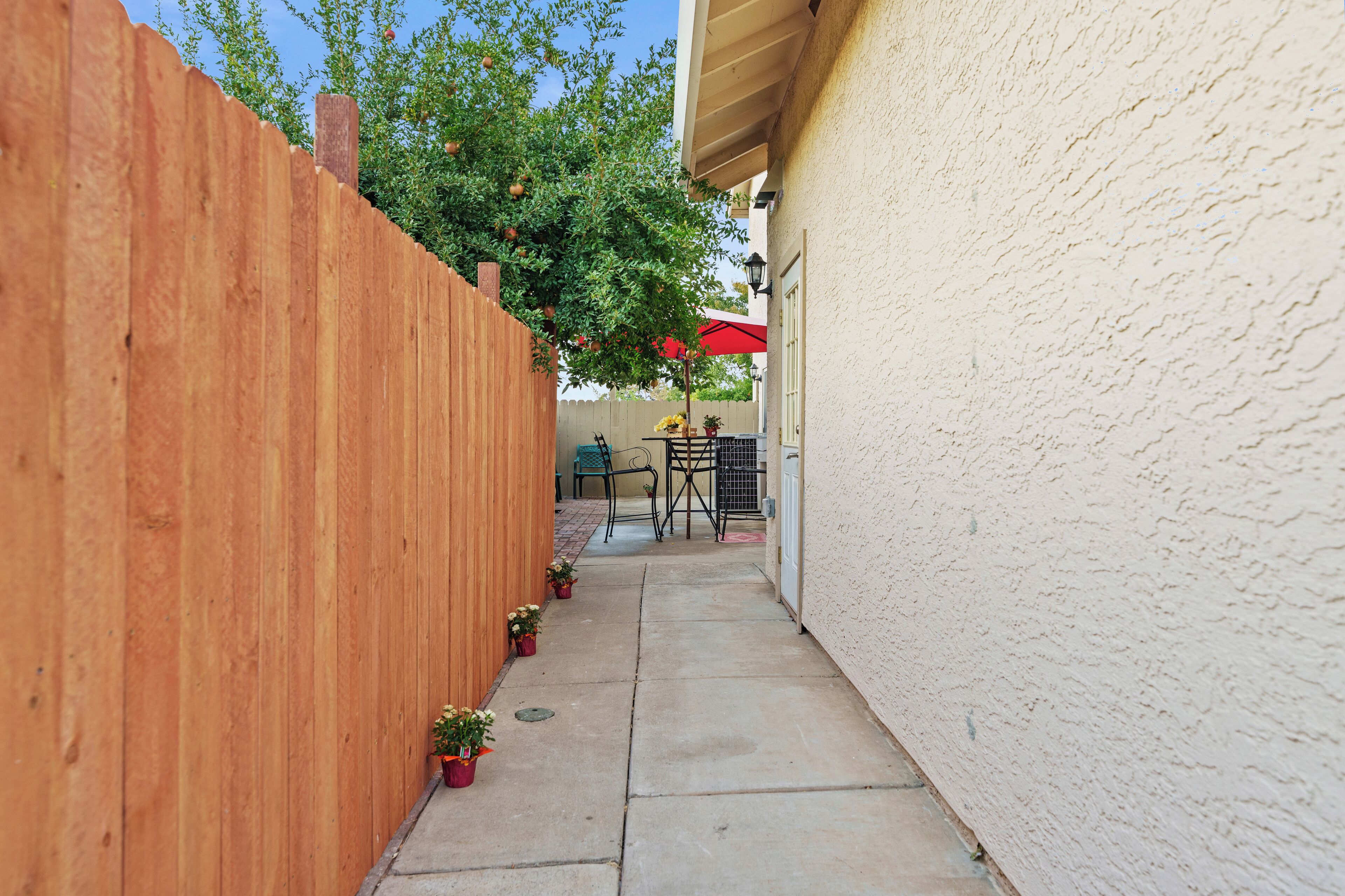 A narrow concrete pathway flanked by a wooden fence and a textured wall, leading to a patio area with a table and chairs, and potted plants.