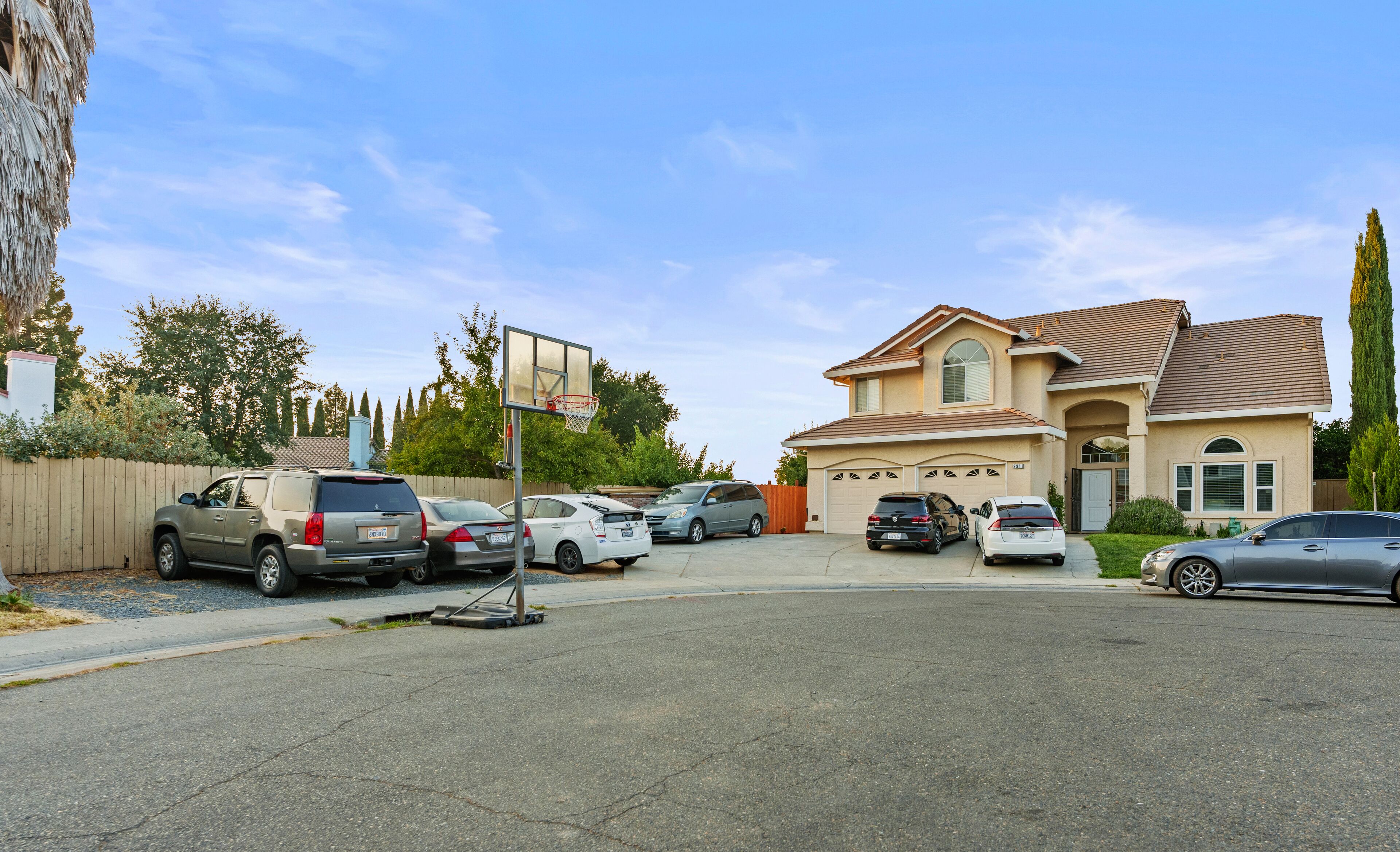 A residential area with a two-story house, a basketball hoop, and several parked cars.