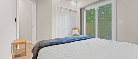 A bedroom with a large bed, a wooden stool, a white door, and a sliding glass door leading to a balcony.