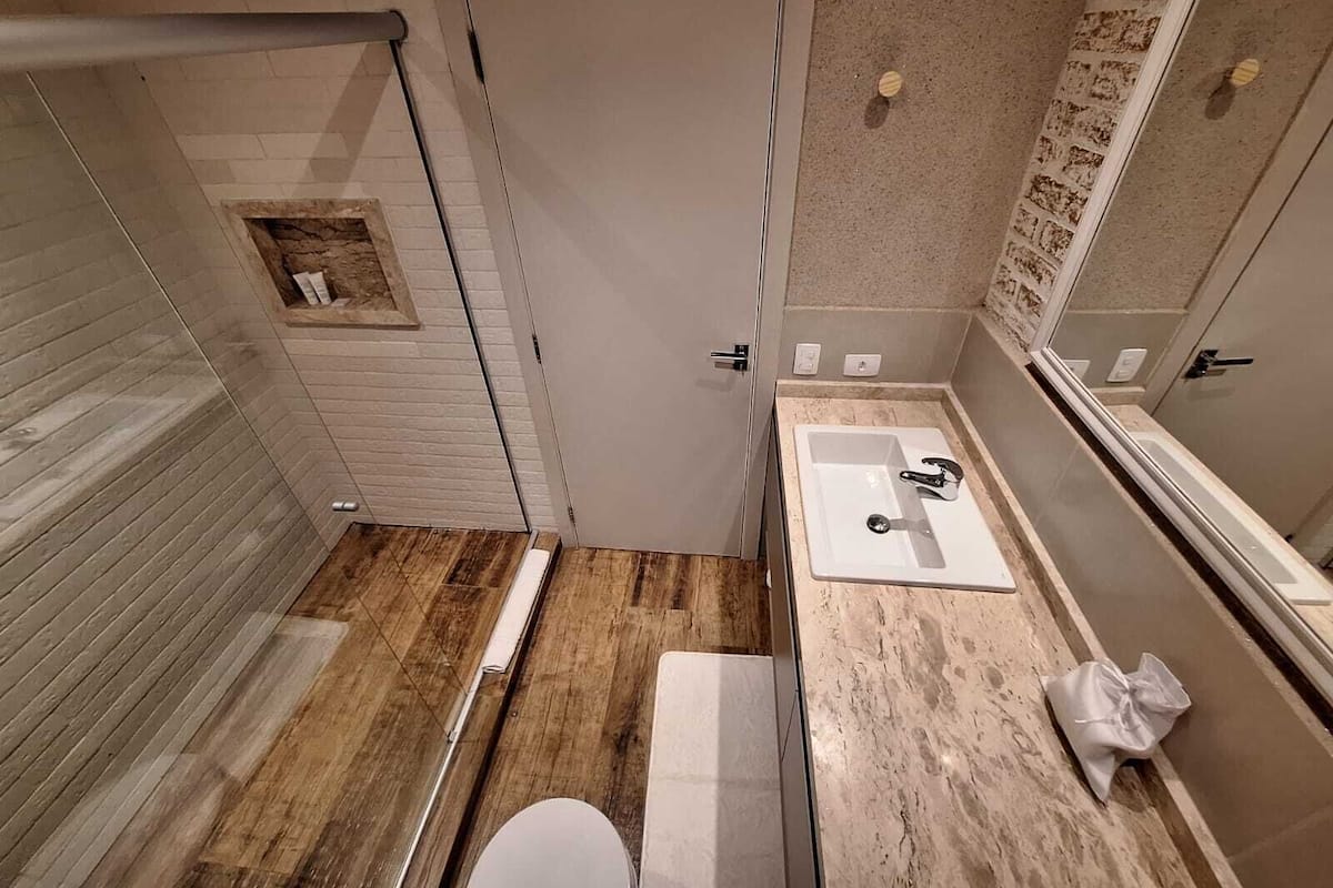 Un baño con piso de madera, inodoro blanco, lavamanos con espejo y zona de ducha.