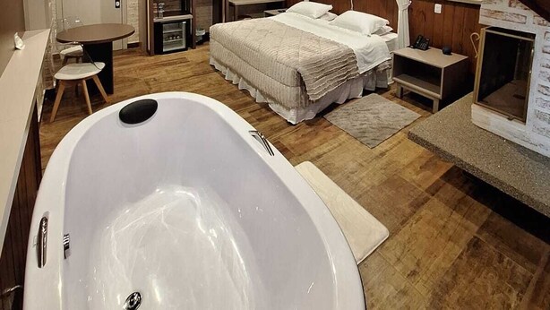 A bedroom with a large bathtub, a bed with white bedding, a wooden ceiling, and a small desk with a chair. - Château Suisse Boutique Hotel (Campos do Jordão)