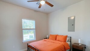 Um quarto com uma cama, uma mesinha de cabeceira, um ventilador de teto, uma janela e um ponto de luz fixado na parede.